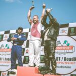 Three racers celebrate on the podium after a motorsport event, holding trophies.